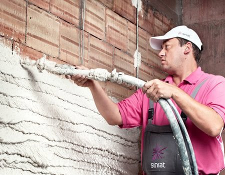 Slika radnika u sivim hlačama sa natpisom Siniat koji pomoću stroja za žbukanje nanosi žbuku na zid od cigle.